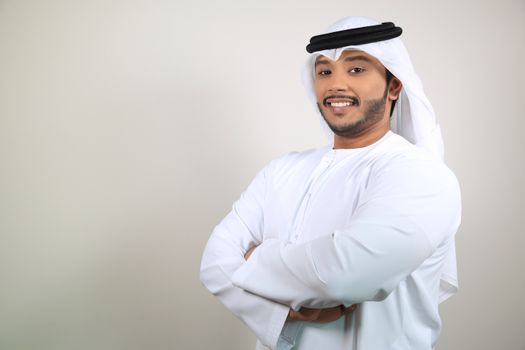 Smiling Middle Eastern man in a white kandura crossing his arms, exuding confidence indoors.