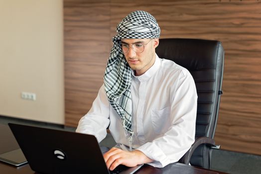 Middle Eastern man in traditional attire working on a laptop in an office setting.